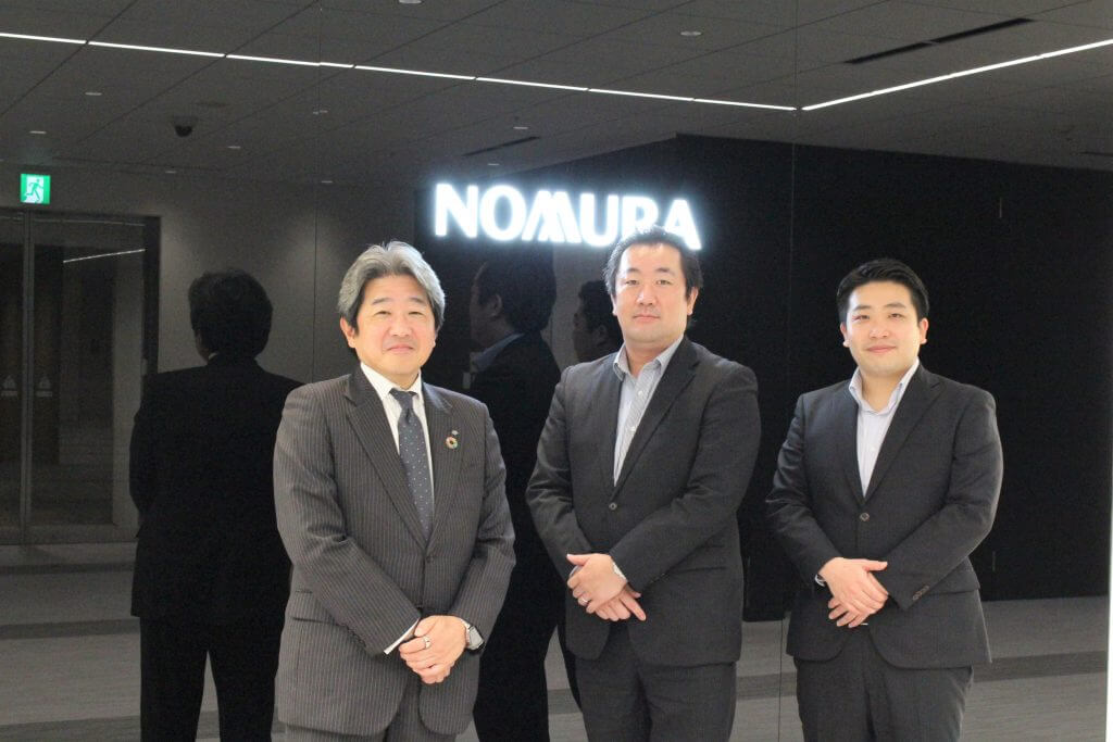 NOMURAの文字をバックに立つ3人のビジネスマン