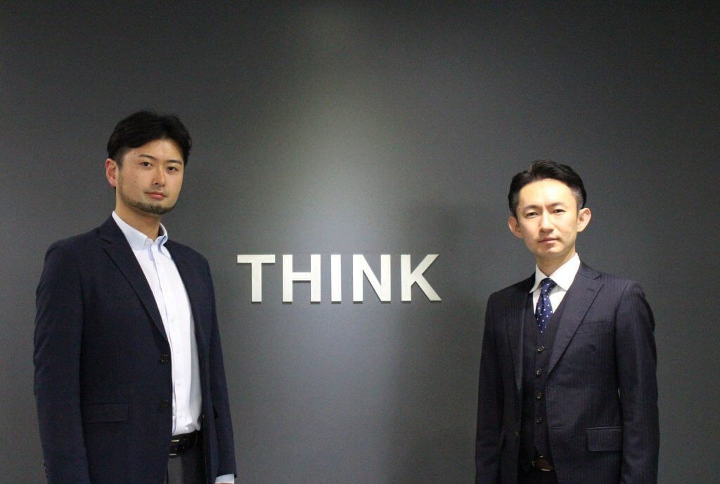 THINKの文字を背景に立つ男性二人